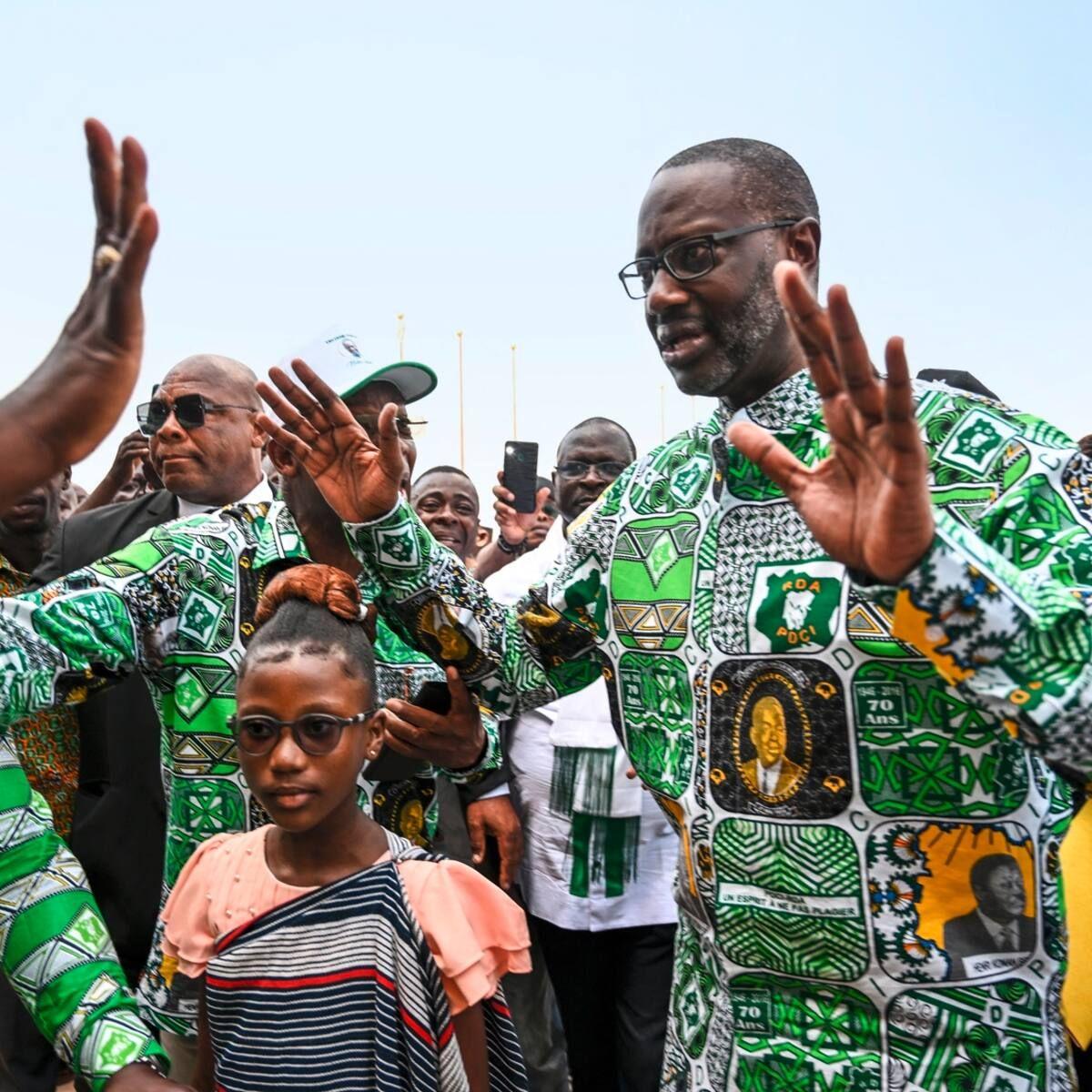 Man in patterned clothing raising hands among a crowd, standing next to a young girl with glasses.