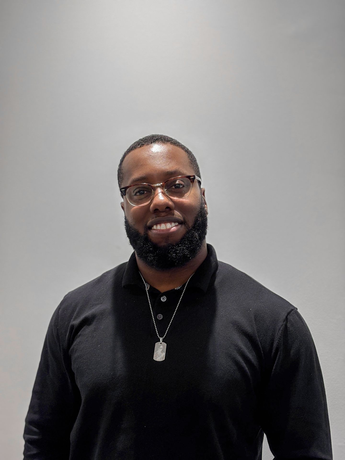 Person wearing a black collared shirt with a dog tag necklace against a gray background.