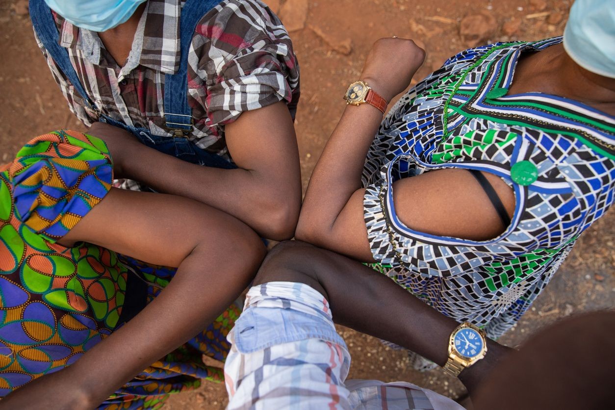 Group of people touching elbows while wearing face masks and colorful clothing, photographed from above.