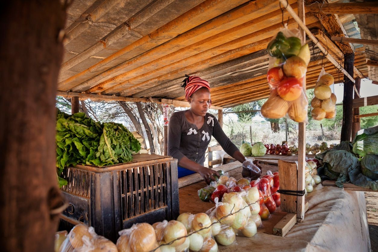 Person arranging fresh vegetables at a market stall under a wooden shelter.